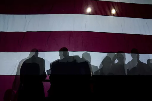 Silhouettes of people are seen on an American flag as President Joe Biden speaks at Max S. Hayes Hight School, Wednesday, July 6, 2022, in Cleveland. Conspiracy theories are nothing new. Humans have always speculated about secret motives and plots as a way to understand their world and avoid danger. But these days, conspiracy theories and those who believe them seem to be playing an outsized role in our politics and culture. (AP Photo/Evan Vucci, File)