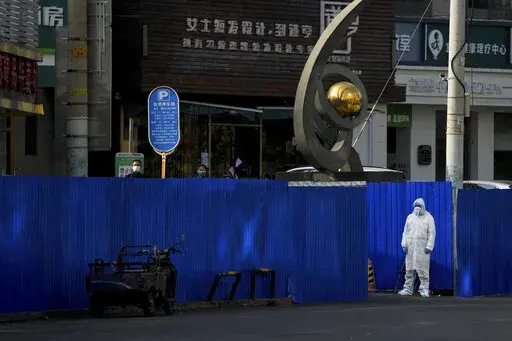 A worker in protective clothing keeps watch as residents wearing face masks stand near metal barriers set up around shuttered shop houses that were locked down as part of COVID-19 controls in Beijing on Thursday, Nov. 10, 2022. China's capital Beijing has closed city parks and imposed other restrictions as the country faces a new wave of COVID-19 cases. (AP Photo/Andy Wong)