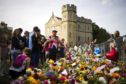 People gather at flowers and messages to tribute Queen Elizabeth II, in front of Windsor Castle in Windsor, England, Sunday, Sept. 11, 2022. Queen Elizabeth II, Britain's longest-reigning monarch and a rock of stability across much of a turbulent century, died Thursday Sept. 8, 2022, after 70 years on the throne. She was 96. (AP Photo/Markus Schreiber)